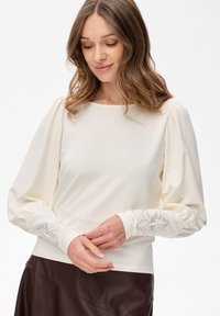 Frau mit welligem braunem Haar, die ein cremefarbenes Langarmoberteil mit bestickten Manschetten und einen dunkelbraunen Lederrock trägt. Sie schaut nach unten und hat die Hände gefaltet.