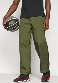 Ελαφριά παντελόνια σε ελιά πράσινο χρώμα με τσαλακωτή υφή. Συνδυασμένα με αθλητικά παπούτσια σε μαύρο και κόκκινο χρώμα και κρατώντας μια μαύρη μπάλα μπάσκετ με κόκκινες λεπτομέρειες.