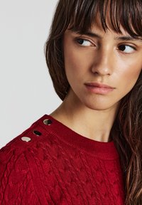 Frau mit langen braunen Haaren, die einen roten Kabelstrickpullover mit drei silbernen Knöpfen an der Schulter trägt und nach rechts schaut.