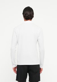 Man with dark hair stands facing away, wearing a white long-sleeve polo shirt and black shorts against a plain white background.