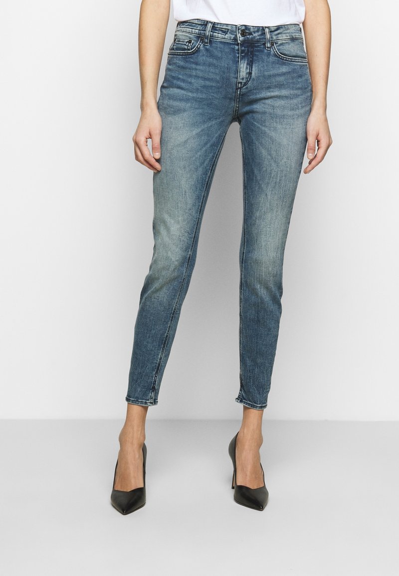 Una persona que lleva jeans ajustados azul y tacones altos negros está de pie contra un fondo liso, con las manos en las caderas.