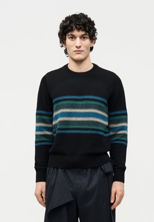 Ung person med krøllet hår iført en sort sweater med grønne, blå og grå vandrette striber og mørke bukser med tynde striber.