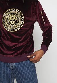 Felpa in velluto borgogna con emblema di leone dorato, design rotondo, polsini e orlo a costine, e una tasca con zip sulla manica sinistra.