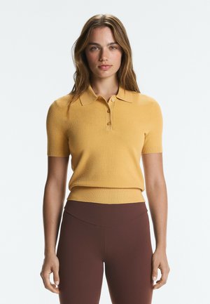 Femme aux longs cheveux bruns portant un polo jaune moutarde à manches courtes et un pantalon marron taille haute, debout sur un fond uni.