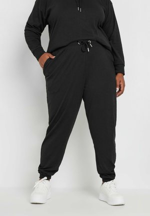 Schwarzes, geripptes Lounge-Set mit Kapuzenpullover und Jogginghose. Mit einem Kordelzugbund und Seitentaschen ausgestattet. Kombiniert mit weißen Turnschuhen.