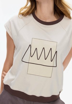 Frau trägt ein beige Kurzarmshirt mit dunkelbraunem Kragen und Saum, das auf der Vorderseite ein abstraktes Zickzackmuster über einem hellen Rechteckdesign zeigt.