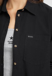Camicia nera con colletto a punta, con taschino frontale e bottoni marroni. Indossata sopra un top grigio con logo.