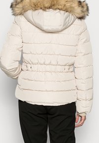 Kvinna iklädd en beige puffjacka med en huva i päls, insnärjd midja och svarta byxor, sedd bakifrån.