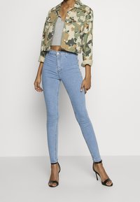 Chaqueta corta con estampado de camuflaje, top de punto gris, jeans de cintura alta azul claro, tacones negros de tiras. Tejido suave, ajustada al cuerpo, diseño casual.