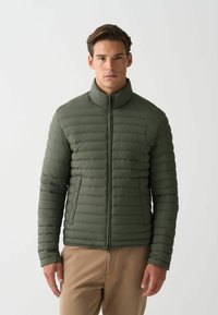 Oliivivärinen puhvijakku, jossa on vaakaompeleet, korkea kaulus, vetoketjusulku ja sivutaskut. Valmistettu kevyestä kankaasta, jossa on sileä pinta.