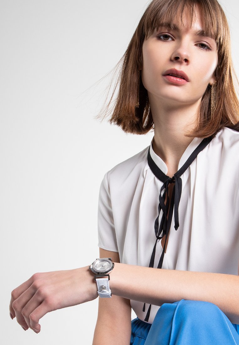 Montre argentée avec un cadran rond et un bracelet en cuir blanc. Le modèle porte une blouse blanche avec un ruban noir et un pantalon bleu.