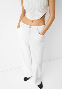 Los pantalones anchos blancos tienen un ajuste relajado, con bolsillos frontales y un sutil acento de botón de metal en la cintura. Textura de tela de algodón suave.