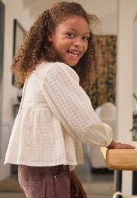 Light, textured white blouse with long sleeves, gathered waist, and a relaxed fit, paired with pink pants. Wooden table in the background.