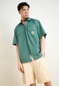 Kortermet skjorte med knapper i grønt stoff med to brystlommer; beige shorts; har et merketikett på skjortelommen.