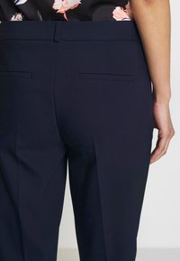 Námořnické kalhoty s hladkou texturou, které mají dvě zadní kapsy a čistý design. Tkanina se zdá být lehká a strukturovaná.