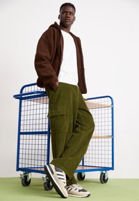 Brun fleece zip-up hoodie, vit t-shirt, mörkgrön manchesterbyxor med fickor och beige sportskor med svarta detaljer.