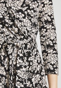 Robe portefeuille fleurie noire et blanche avec manches 3/4, dotée d'une ceinture nouée à la taille et d'un motif floral détaillé.