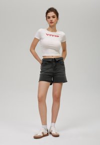 Camiseta corta blanca con el texto rojo "VIVID", combinada con pantalones cortos de mezclilla negros de tiro alto. La modelo lleva zapatillas beiges y blancas con calcetines grises.