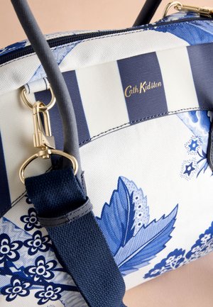 Mėlynos ir baltos spalvos raštuotas krepšys su gėlių motyvais, dryžuotomis dalimis, auksinės spalvos detalėmis, tvirtomis rankenomis ir tekstūruota paviršiaus struktūra. Matomas prekės ženklo logotipas.
