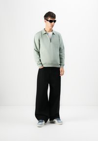 Joven con gafas de sol negras, sudadera verde salvia con cremallera, pantalones anchos negros y zapatillas blancas de pie contra un fondo blanco.