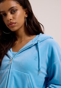 Mladá žena oblečená v svetlomodrej mikine na zips s kapucňou a kovovým zipsom, s veľkými striebornými kruhovými náušnicami.