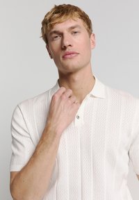 Witte polo met korte mouwen, met een gestructureerd, gebreid patroon en een contrasterende kraag. Inclusief een knop bij de hals en subtiele verticale lijnen.