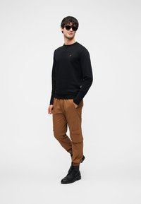 Černá mikina s dlouhými rukávy a malým červeným logem, v kombinaci s hnědými kalhotami typu cargo s bočními kapsami a elastickými manžetami. Černé boty.