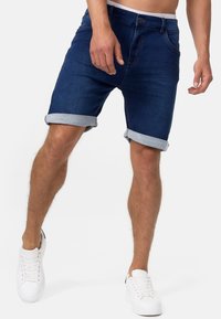 Shorts en denim bleu foncé avec des ourlets roulés, présentant un design classique, cinq poches et une fermeture à bouton. Portés avec des baskets blanches.