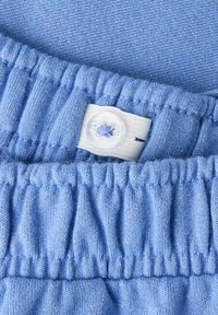 Gaiši zils audums ar tekstūru un maigu spīdumu, kam ir sapludināts elastīgs jostasvieta un neliels balts pogas uzraksts ar ziedu dizainu.