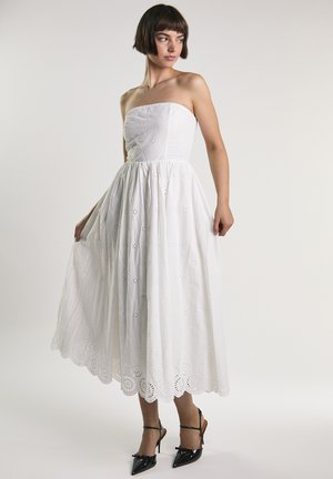 Kobieta w białej, bezramkowej sukience midi z ażurowym wzorem i czarnych szpilkach, pozująca na jednolitym jasnym tle.