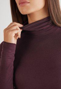 Burgundské rolákové triko s přiléhavými dlouhými rukávy, hladká látka a vrstvený detail výstřihu. Pohled zblízka na krk a ruku.