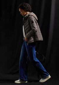Veste matelassée gris foncé avec capuche, associée à un jean bleu à jambe large et des baskets claires, sur fond noir.