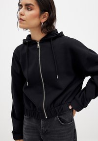 Black cropped hoodie with a silver zipper, adjustable drawstring, and elastic waist. Features long sleeves and a smooth fabric texture.