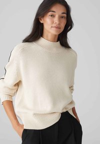 Cremfarbener gestrickter Pullover mit hohem Rippkragen, lässiger Passform und schwarzen Akzentstreifen an den Ärmeln. Rippbündchen an Saum und Ärmeln.