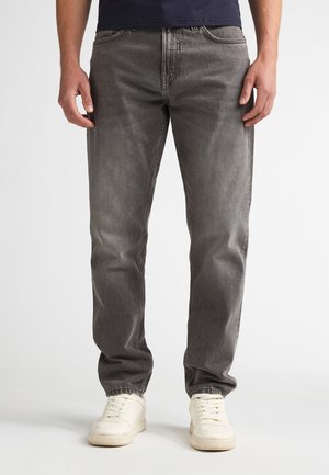 Man wearing faded gray jeans and white sneakers standing against a plain white background.