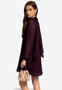 Paarse chiffon jurk met lange, vloeiende mouwen en een hoge hals, gecombineerd met een kleine beige satijnen handtas met een kettinghengsel.