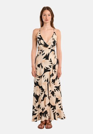 Donna in piedi che indossa un abito lungo nero con grandi motivi a foglia beige, spalline sottili e sandali piatti marroni.