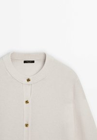 Vaaleanbeige neuletakki, jossa on pyöreä kaula-aukko, ribbireunat ja neljä kultasävyistä napin yksityiskohtaa etuosassa, pehmeällä neulotulla pinnalla.