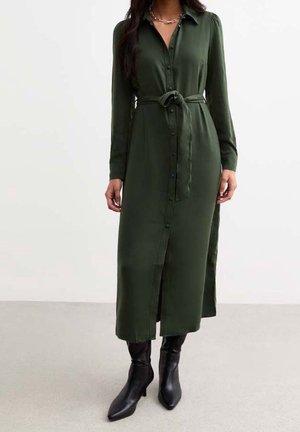 Femme portant une robe midi verte foncée à manches longues avec boutons, une ceinture nouée à la taille et des bottines noires à talons.