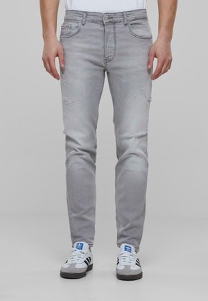 Jeans de mezclilla gris claro con corte ajustado. Presentan detalles desgastados y cinco bolsillos. Combinados con zapatillas deportivas blancas y grises.