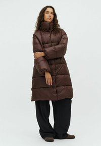Cappotto puffo marrone con colletto alto, caratterizzato da un design trapuntato e maniche ampie. Indossato sopra pantaloni larghi scuri e scarpe marroni.