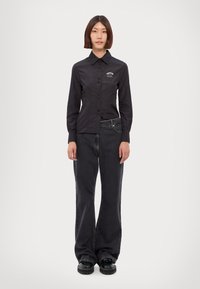 Camicia nera a maniche lunghe con bottoni, pieghe frontali, colletto slim e logo ricamato, abbinata a jeans scuri dal taglio ampio e scarpe platform nere.