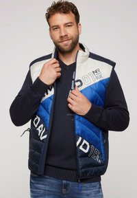 Gilet imbottito blu e bianco con chiusura a zip, caratterizzato da blocchi di colore diagonali e accenti di testo stampati. Indossato sopra un maglione scuro.