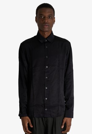 Giovane uomo che indossa una camicia nera a maniche lunghe con bottoni, in piedi davanti a uno sfondo chiaro e semplice, rivolto in avanti con un'espressione neutra.