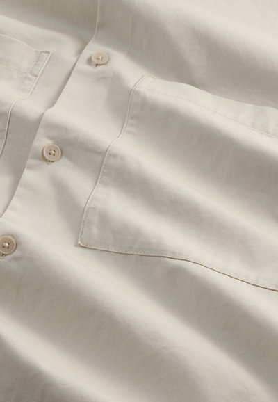 Lys beige skjorte lavet af tekstureret stof, med en brystlomme og knapper i en matchende farve. Glat finish med minimale rynker.
