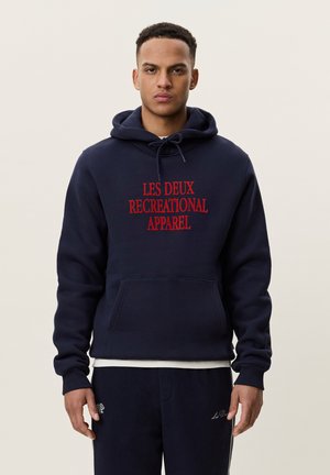 Jeune homme portant un sweat à capuche bleu foncé avec un texte rouge "Les Deux Recreational Apparel" et un pantalon bleu foncé assorti avec des détails brodés blancs.