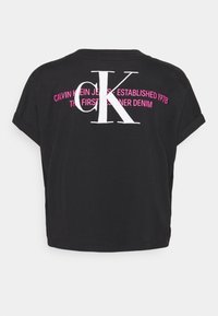 Camiseta negra corta con mangas enrolladas, gran logo blanco "CK" y texto rosa "CALVIN KLEIN JEANS - ESTABLECIDO 1978 THE FIRST CORNER DENIM" en la parte posterior.