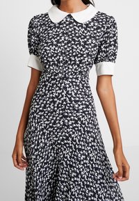 Robe noire avec motif d'oiseaux blancs, manches courtes bouffantes et col blanc à revers. Le tissu présente une texture lisse et un design fluide.