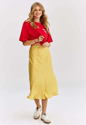 Kobieta uśmiecha się, nosi czerwony krótki t-shirt, żółtą midi spódnicę w wzory oraz białe sportowe buty na jednolitym białym tle.