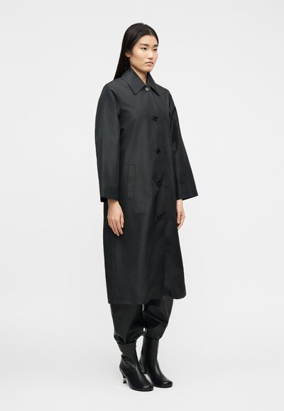 Γυναίκα που φοράει μαύρο μακρύ παλτό με κουμπιά πάνω από μαύρο παντελόνι και μαύρες μπότες με τακούνι, στέκεται μπροστά σε ένα απλό φωτεινό φόντο.